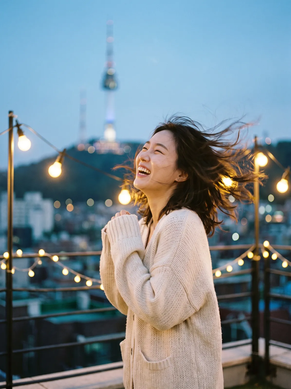 Candid portrait of a Korean woman in her late twenties laughing on a Seoul rooftop at blue hour, oversized cream knit cardigan, wind lifting loose strands of hair, Namsan Tower softly out of focus beh