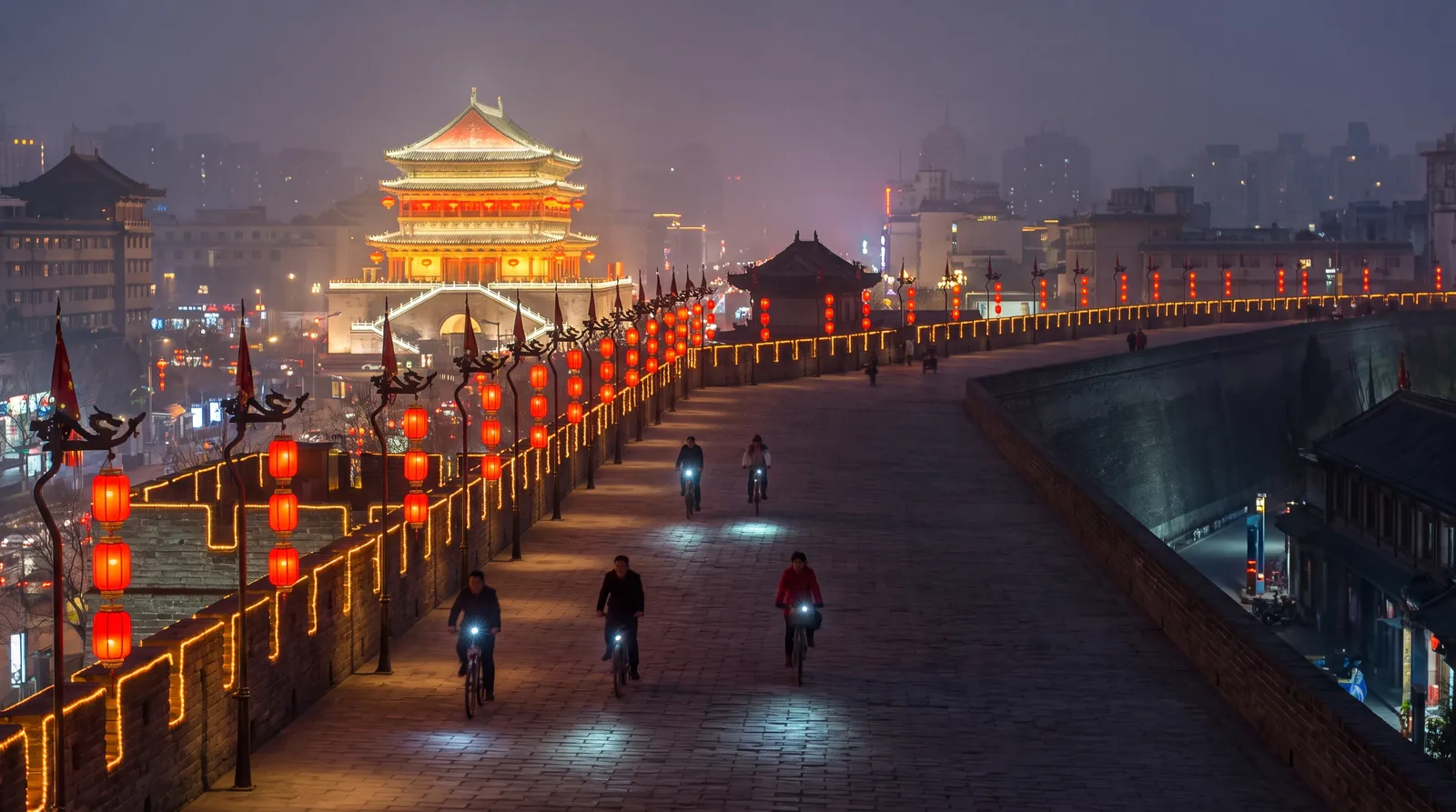 Xi'an city wall at night illuminated with lanterns, cyclists riding on top, the Bell Tower glowing in the distance