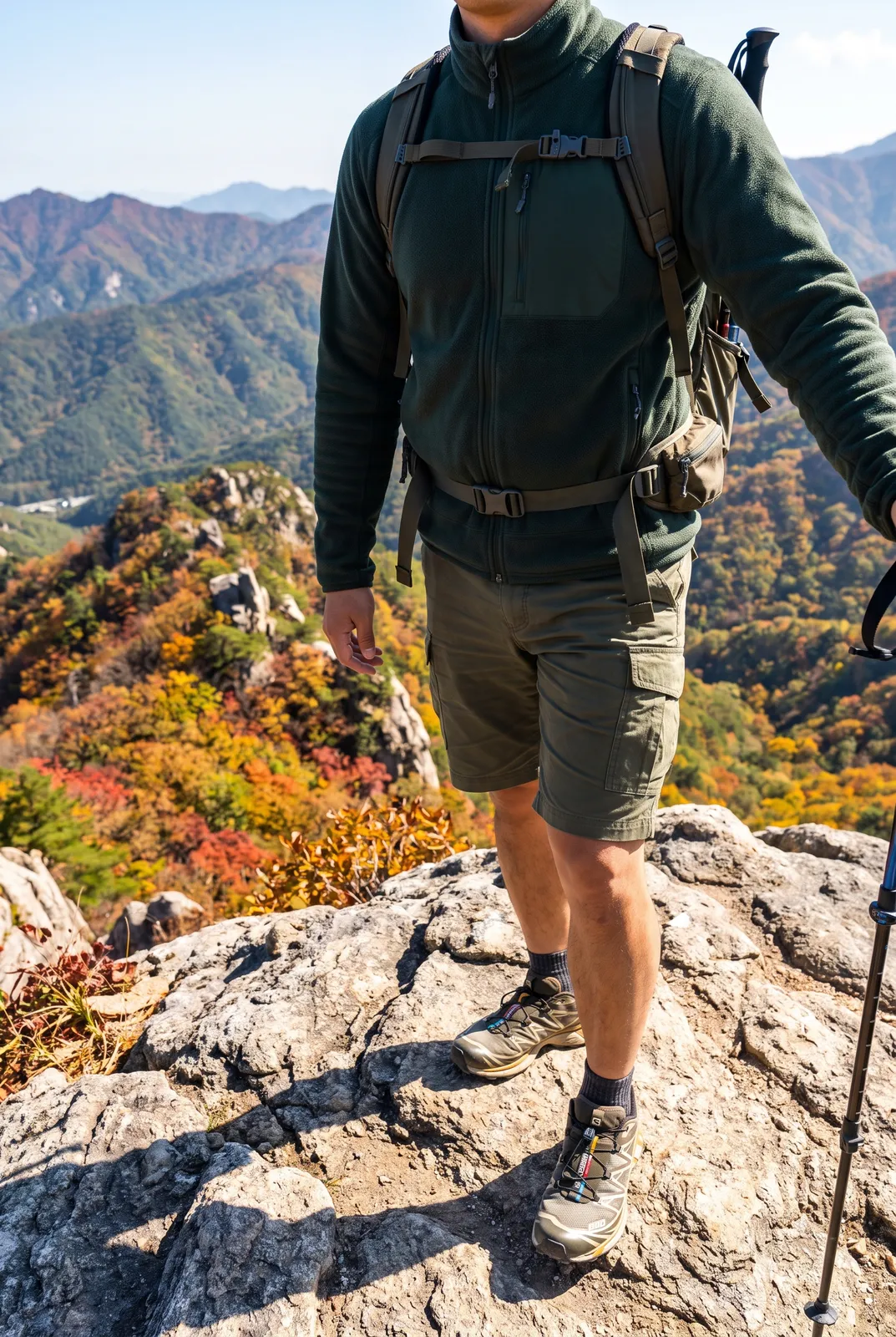 Outdoor gorpcore fashion in the Korean mountains: a model in a technical fleece, cargo shorts and trail shoes standing on a rocky ridge at Seoraksan, autumn colors, bright clear light