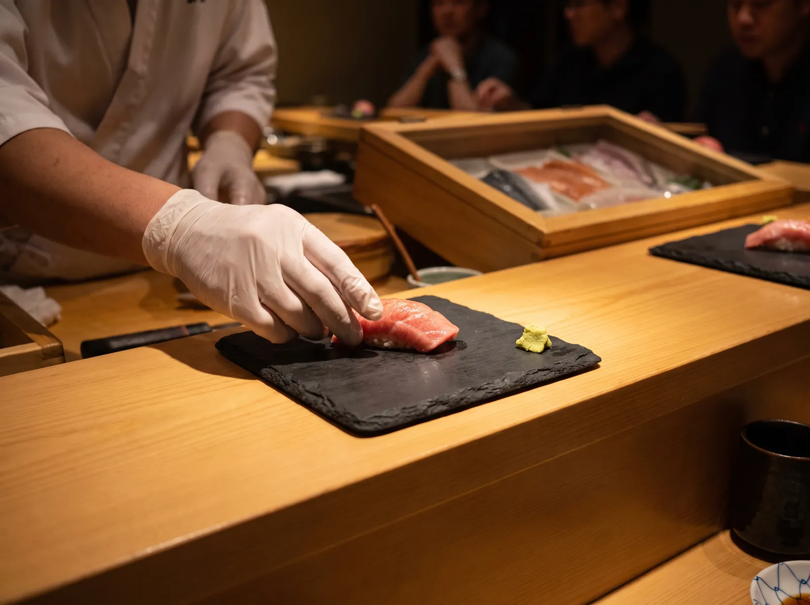 Japanese omakase sushi counter, a chef's hands placing otoro nigiri on a black slate, glossy fish, soft warm spotlight, premium dining