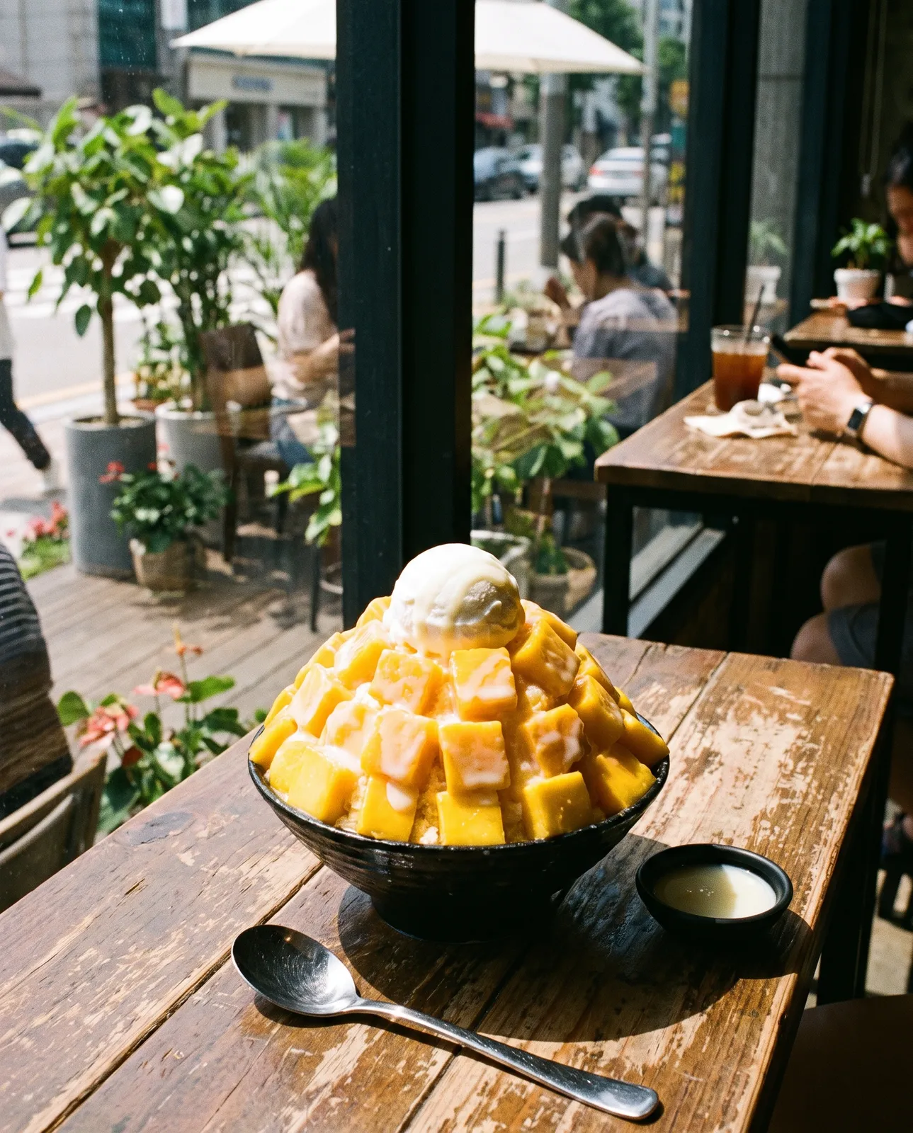 Korean mango bingsu piled high with mango cubes, condensed milk and a scoop of yogurt ice cream on a summer café table by the window, bright fresh light