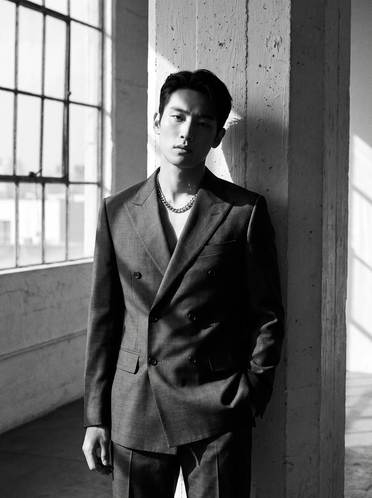 Monochrome fashion portrait of a Korean male model in a tailored charcoal double-breasted suit with no shirt, silver chain necklace, leaning on a concrete column, hard window light, high contrast