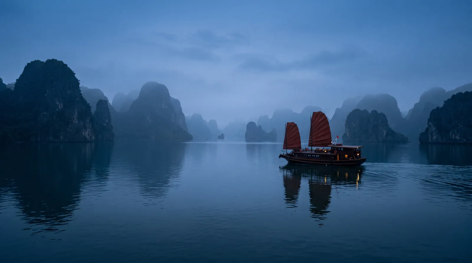 Ha Long Bay at blue hour, limestone islands fading into mist, a junk boat with rust-red sails gliding over calm water, serene landscape photography