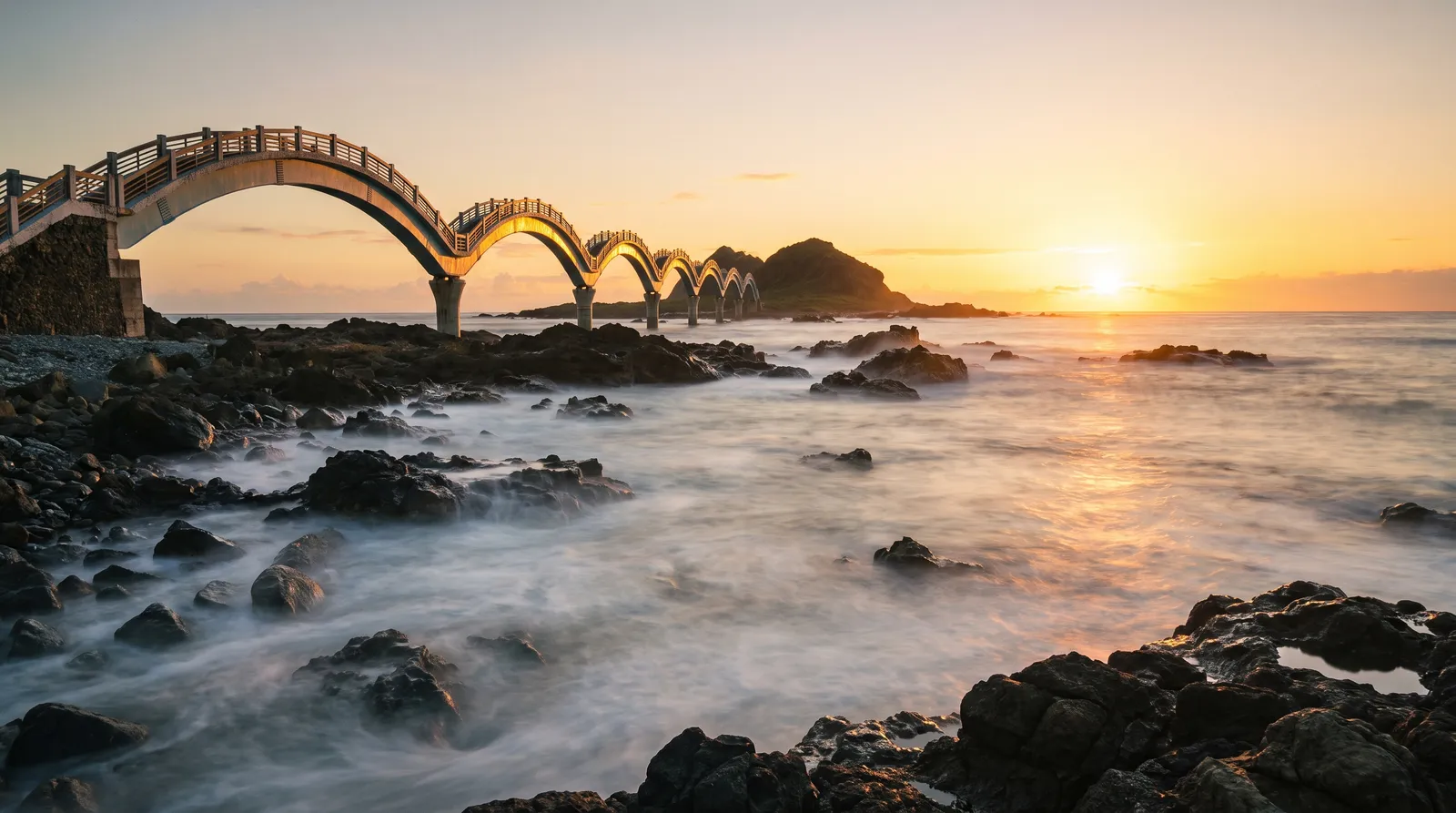 Sanxiantai on Taiwan's east coast at sunrise, a long multi-arched footbridge curving out to a rocky islet, Pacific waves breaking over volcanic rocks, warm gold light, silky long-exposure water