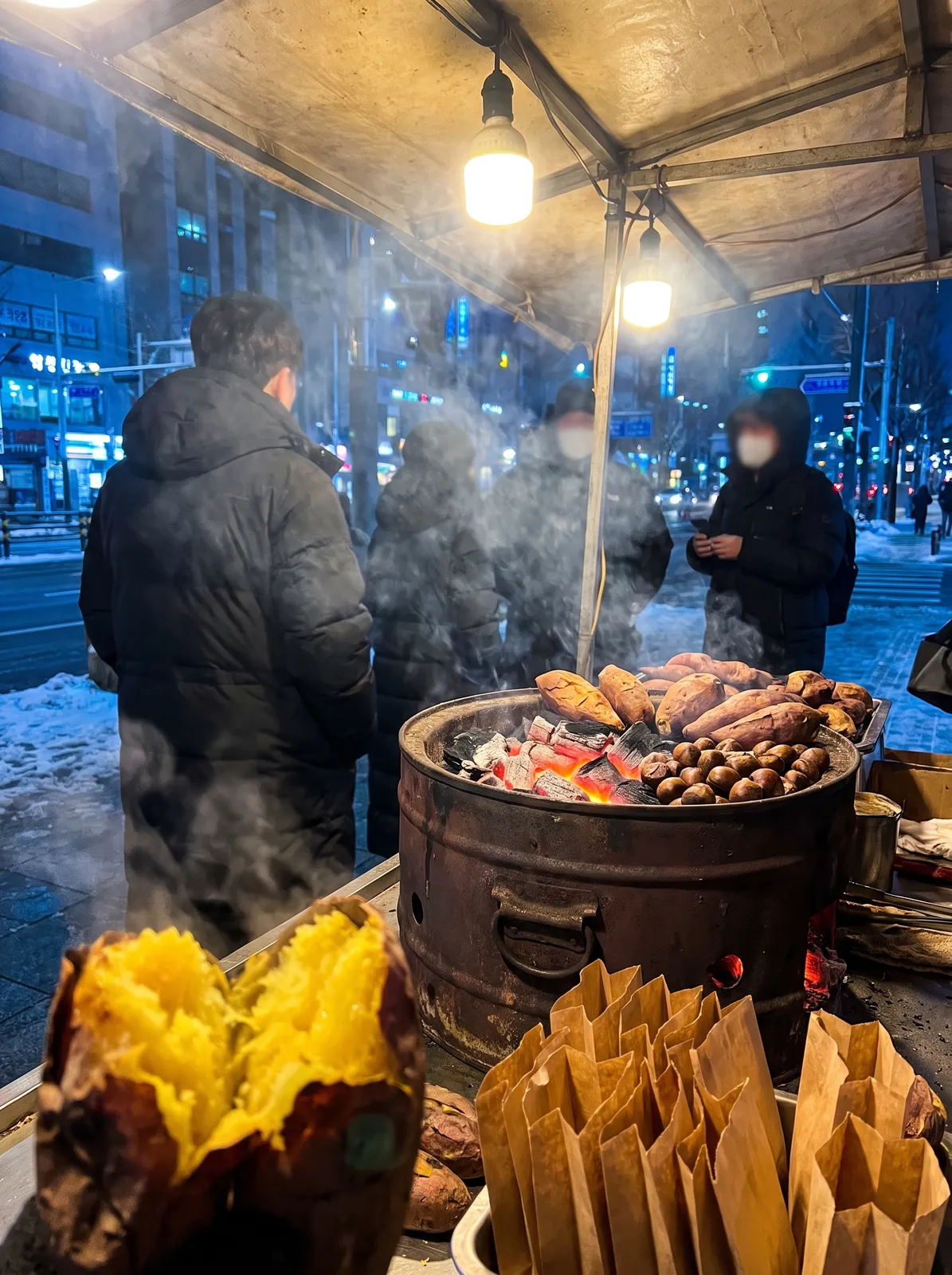 Food photography of roasted sweet potatoes and chestnuts in a charcoal drum at a Seoul winter street stall, one potato split open to golden flesh, paper bags waiting, wisps of smoke…