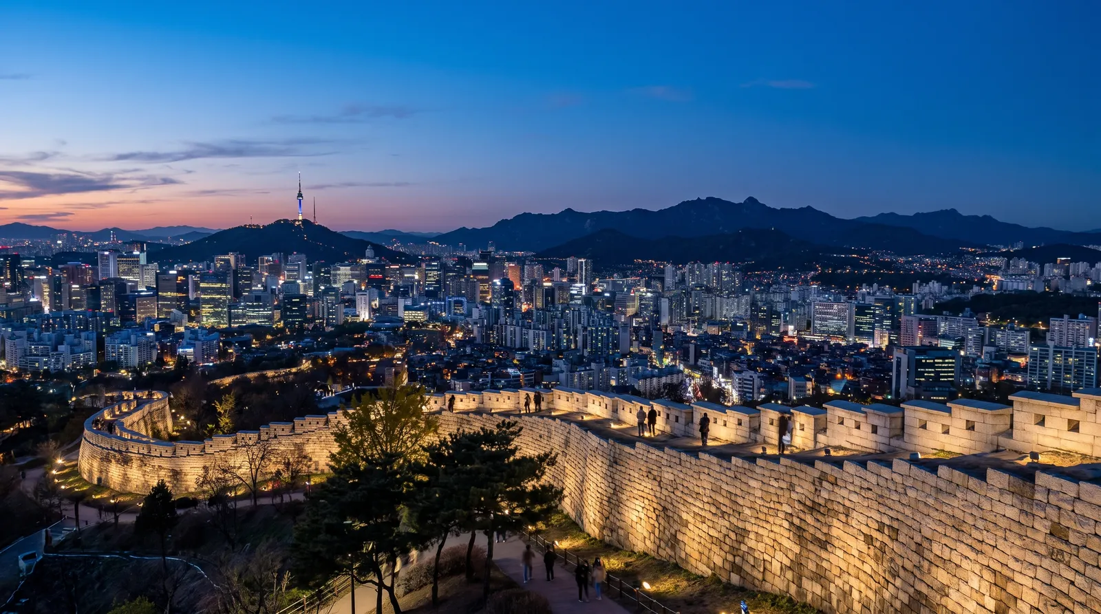 Seoul skyline from Naksan park at blue hour, the old fortress wall in the foreground, dense city lights stretching to the mountains, clear crisp air