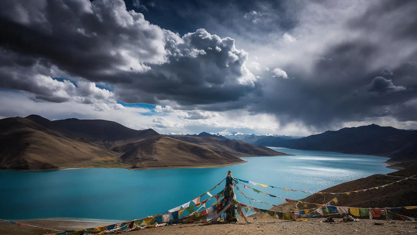 Tibetan plateau with turquoise lake Yamdrok under dramatic clouds, snow mountains on the horizon, prayer flags in the foreground, vast and spiritual