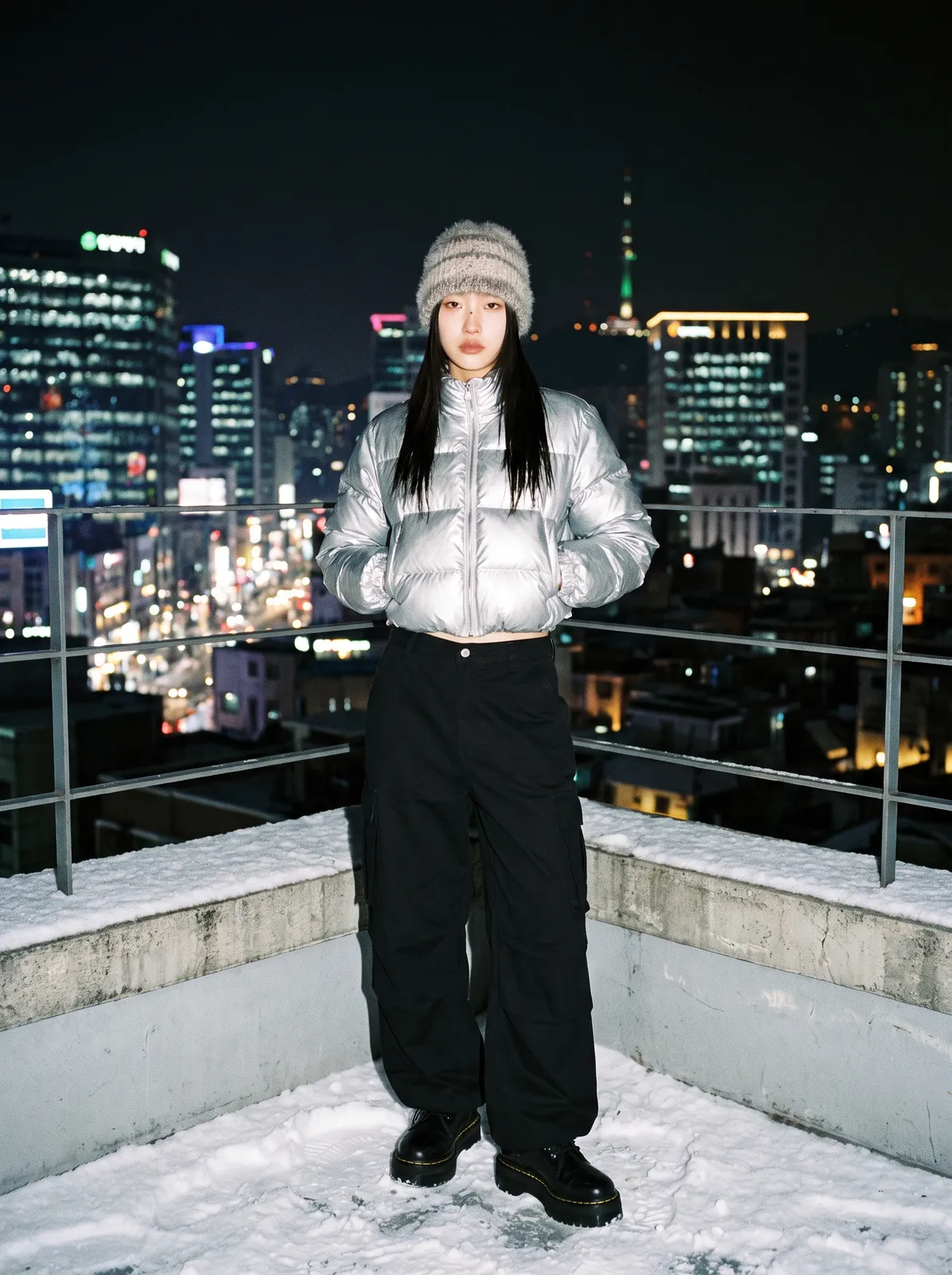 Korean model in a puffy cropped silver jacket and wide black trousers on a snowy Seoul rooftop at night, city lights below, flash photography, Y2K-inspired winter editorial