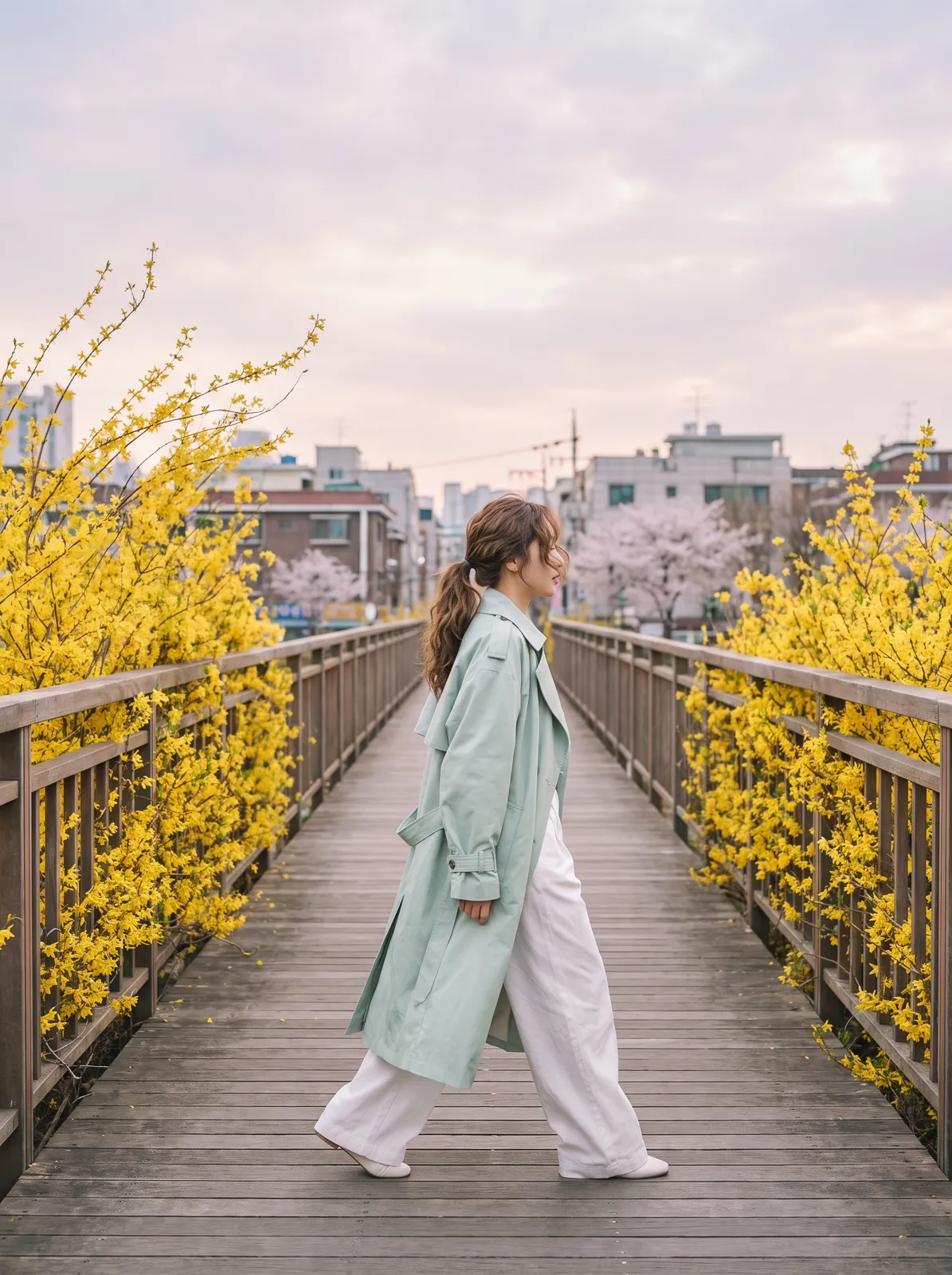Spring fashion editorial of a model in a pale mint trench coat and wide white trousers crossing a Seoul pedestrian bridge lined with blooming forsythia, soft breeze, clean pastel palette