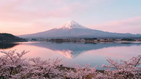 Mount Fuji reflected in Lake Kawaguchi at dawn with cherry blossoms framing the shore, glassy water…