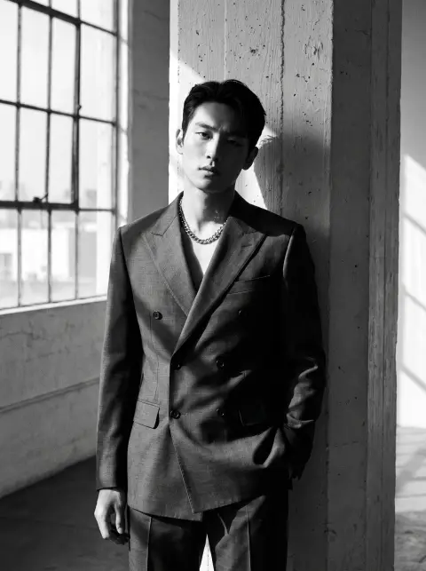 Monochrome fashion portrait of a Korean male model in a tailored charcoal double-breasted suit with no shirt, silver cha