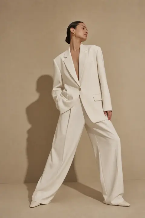 Minimal studio fashion shot of a model in an ivory oversized blazer and wide trousers against a sand-coloured backdrop,