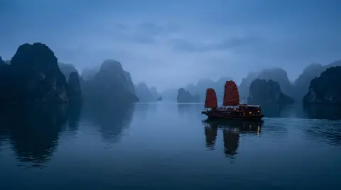 Ha Long Bay at blue hour, limestone islands fading into mist, a junk boat with rust-red sails gliding over calm water…