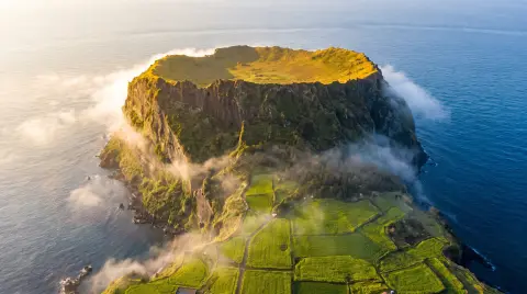 Aerial view of Jeju island's Seongsan Ilchulbong crater at sunrise, rim glowing gold, ocean mist around the base, green