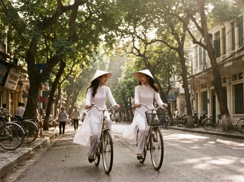 Two Vietnamese sisters in white ao dai cycling side by side along a tree-lined Hanoi street in morning light, sun flicke
