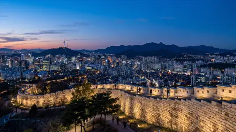 Seoul skyline from Naksan park at blue hour, the old fortress wall in the foreground, dense city lights stretching to th